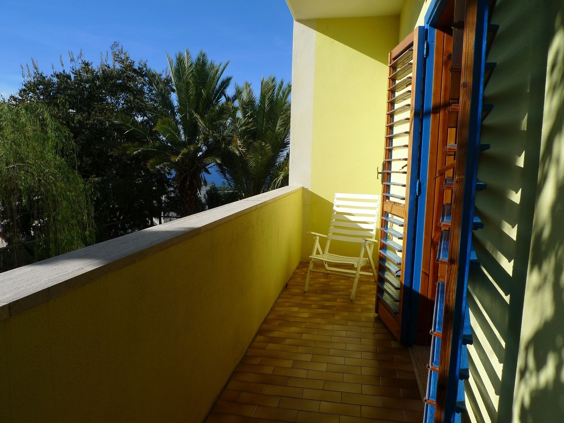 Ein Balkon mit Holzboden, einem weißen Stuhl und einem blau-braunen Metallgitter.