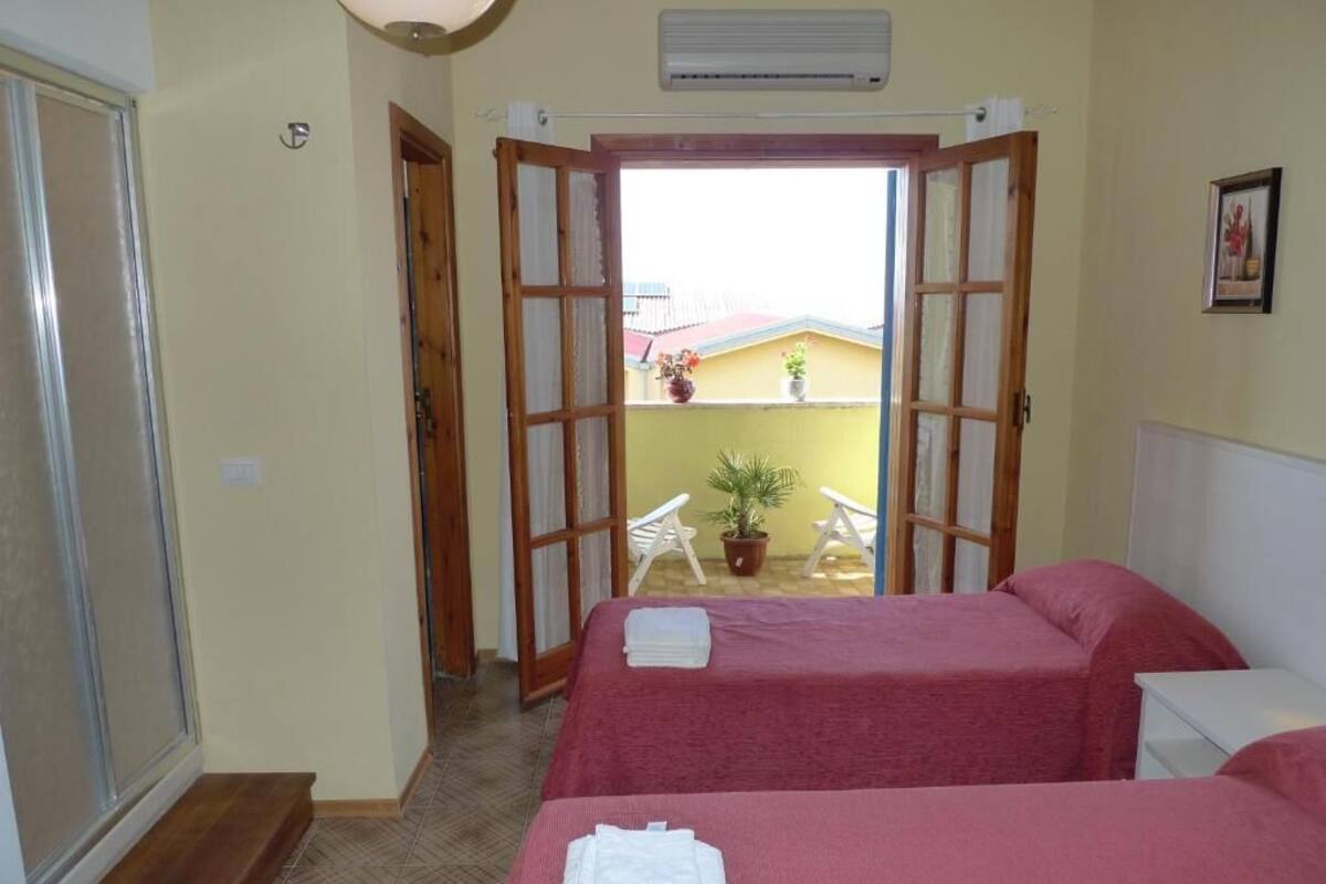 A hotel room with two beds, a balcony with potted plants, and an air conditioning unit.