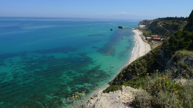 A coastal landscape with clear turquoise waters, a sandy beach, and a rocky cliff with vegetation. - Hotel Alexander (Ricadi)