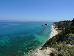 A coastal landscape with clear turquoise waters, a sandy beach, and a rocky cliff with vegetation. - Hotel Alexander (Ricadi)