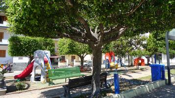 Un terrain de jeux avec un toboggan, des balançoires et un banc à l’ombre d’un arbre.