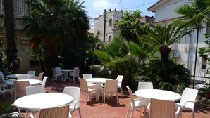 An outdoor patio area with round tables and white wicker chairs, surrounded by palm trees and potted plants. - Hotel Alexander (Ricadi)