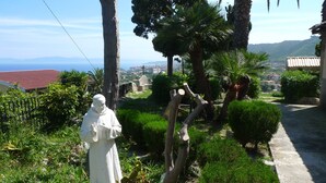 A statue of a robed figure in a garden with a cactus and a sign, surrounded by greenery and a view of a town. - Hotel Alexander (Ricadi)