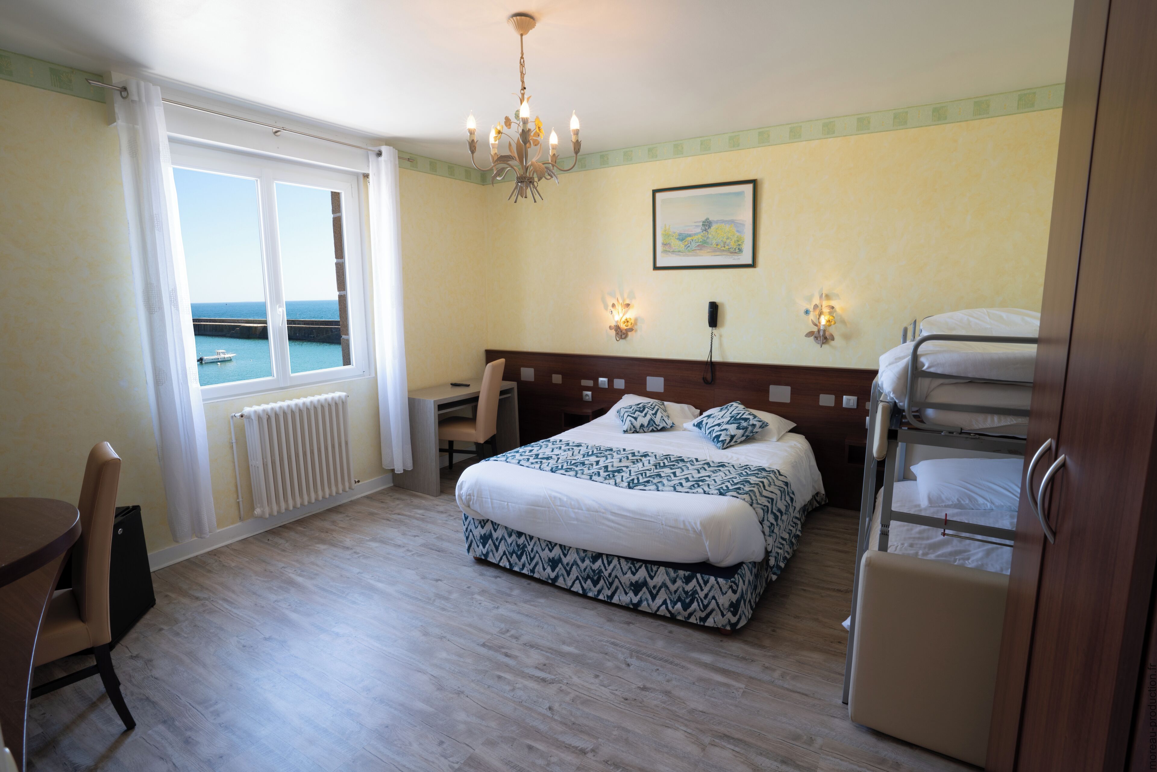 Hotelkamer met een bed, een bureau, een stoel, uitzicht op zee, een kroonluchter en een stapelbed.