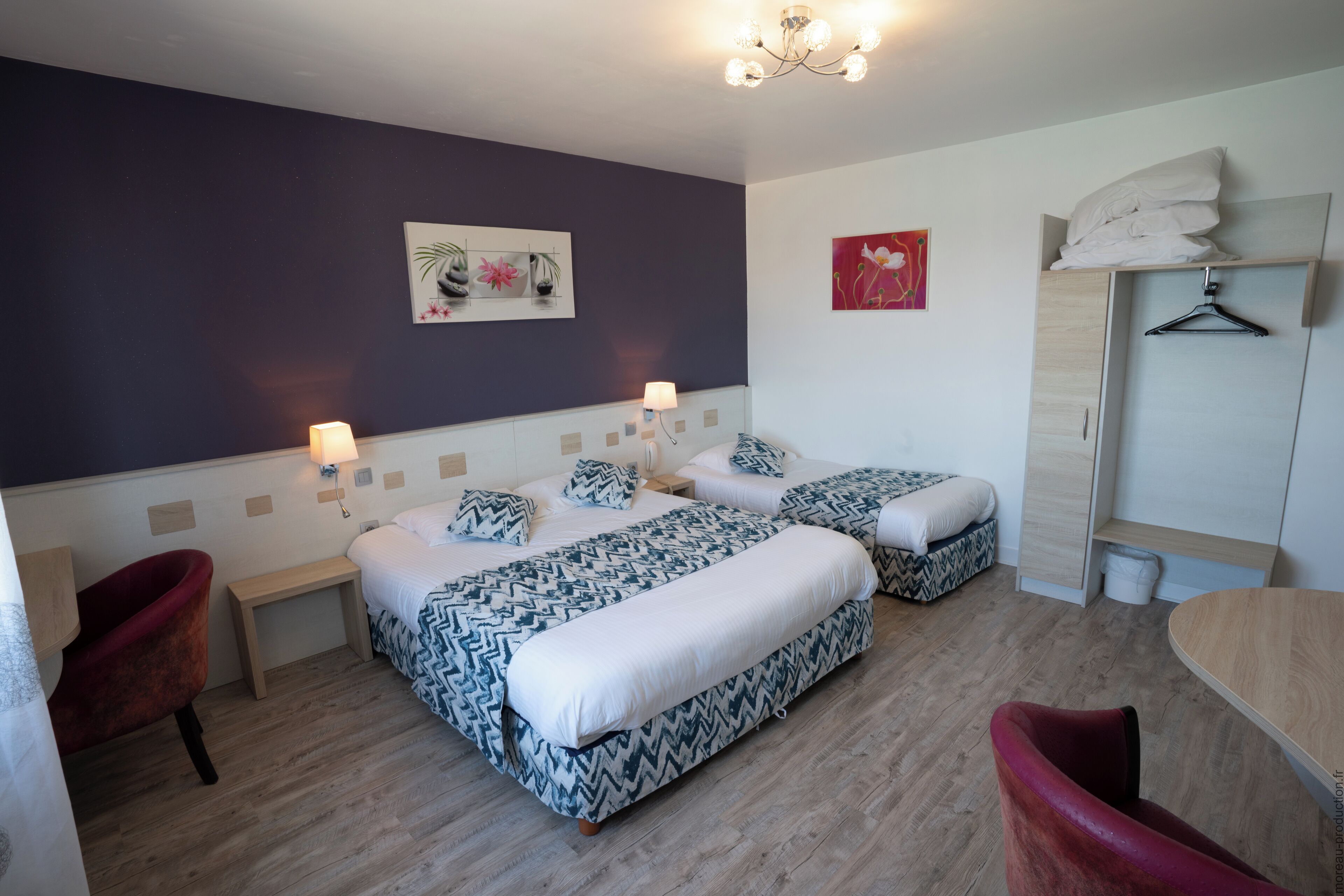 A hotel room with two beds, a wooden floor, a red armchair, a wooden table, and a wardrobe with a hanging shelf.