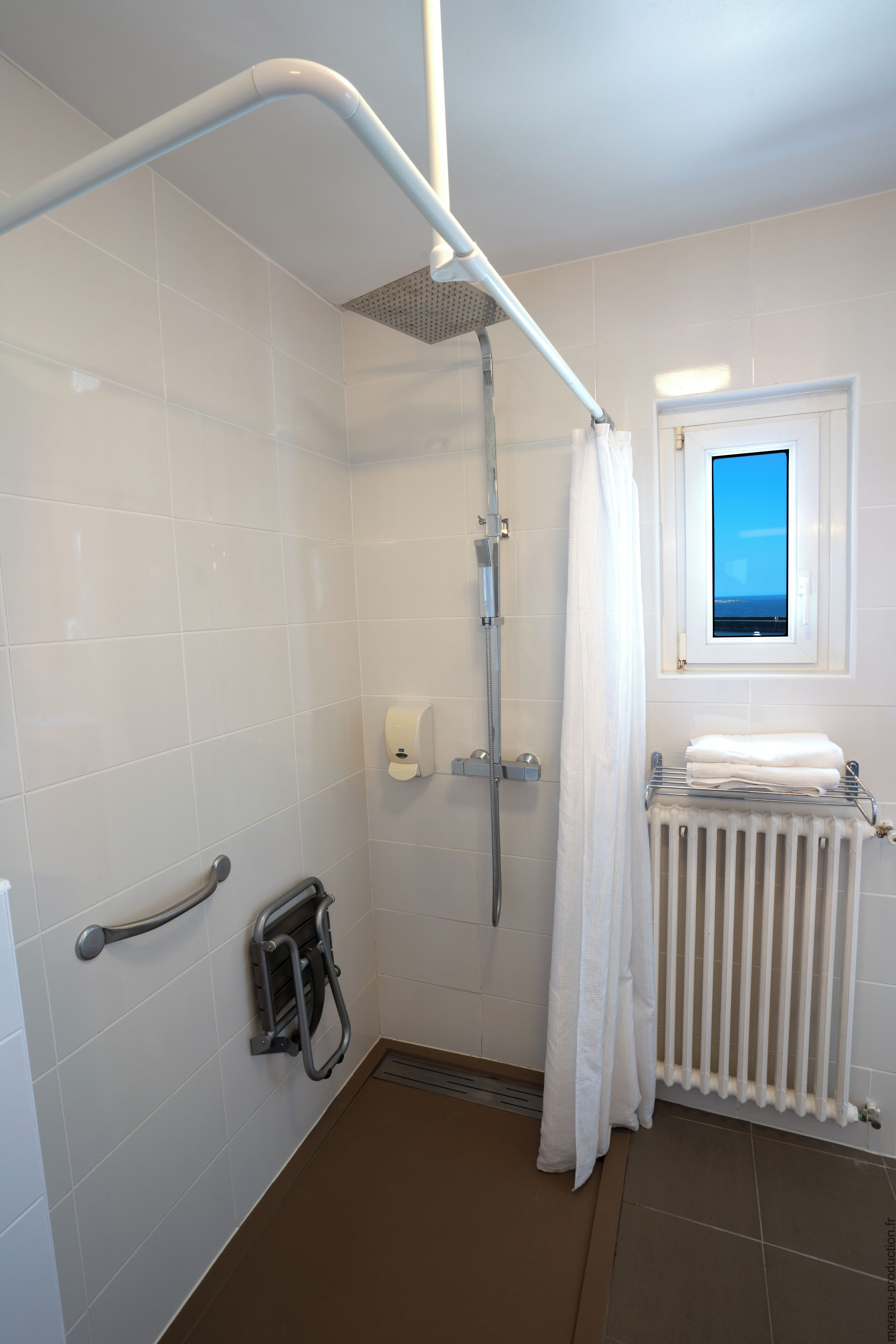 A bathroom with white tiles, a shower with a grab bar, a window with a view, and a radiator.