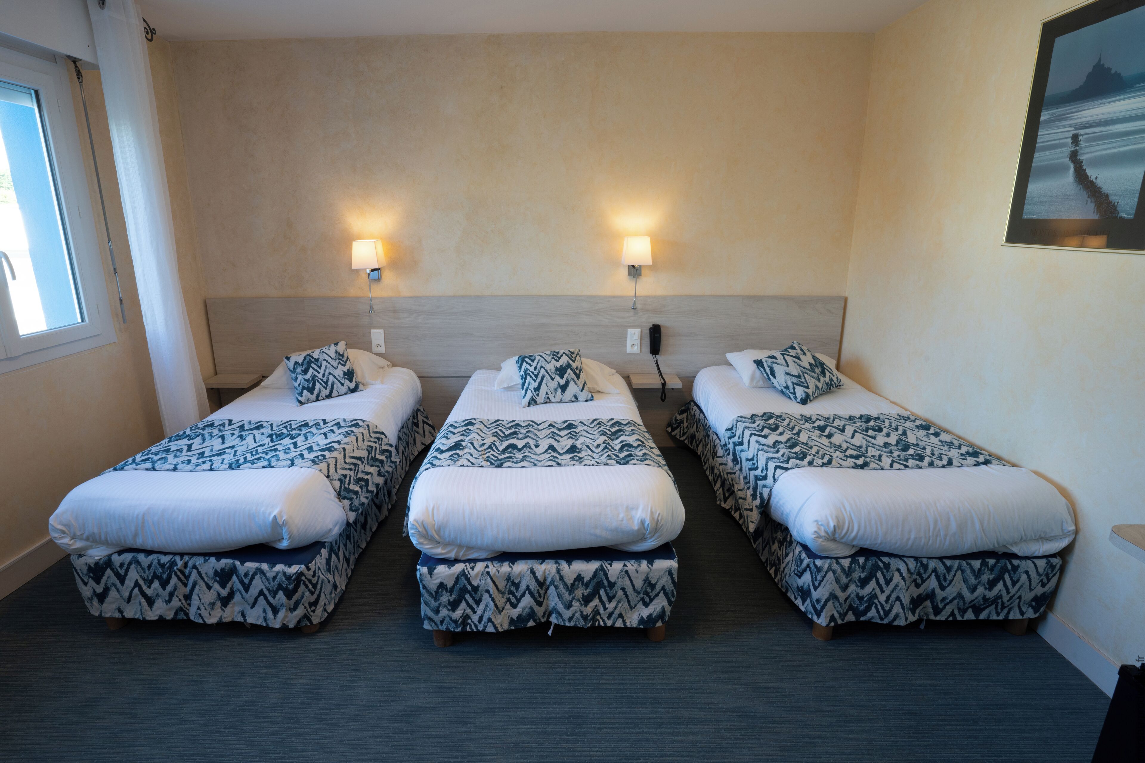 A hotel room with three single beds, each with a patterned bedspread and matching pillows. There are two wall-mounted lamps on either side of the beds, and a framed picture on the wall to the right.