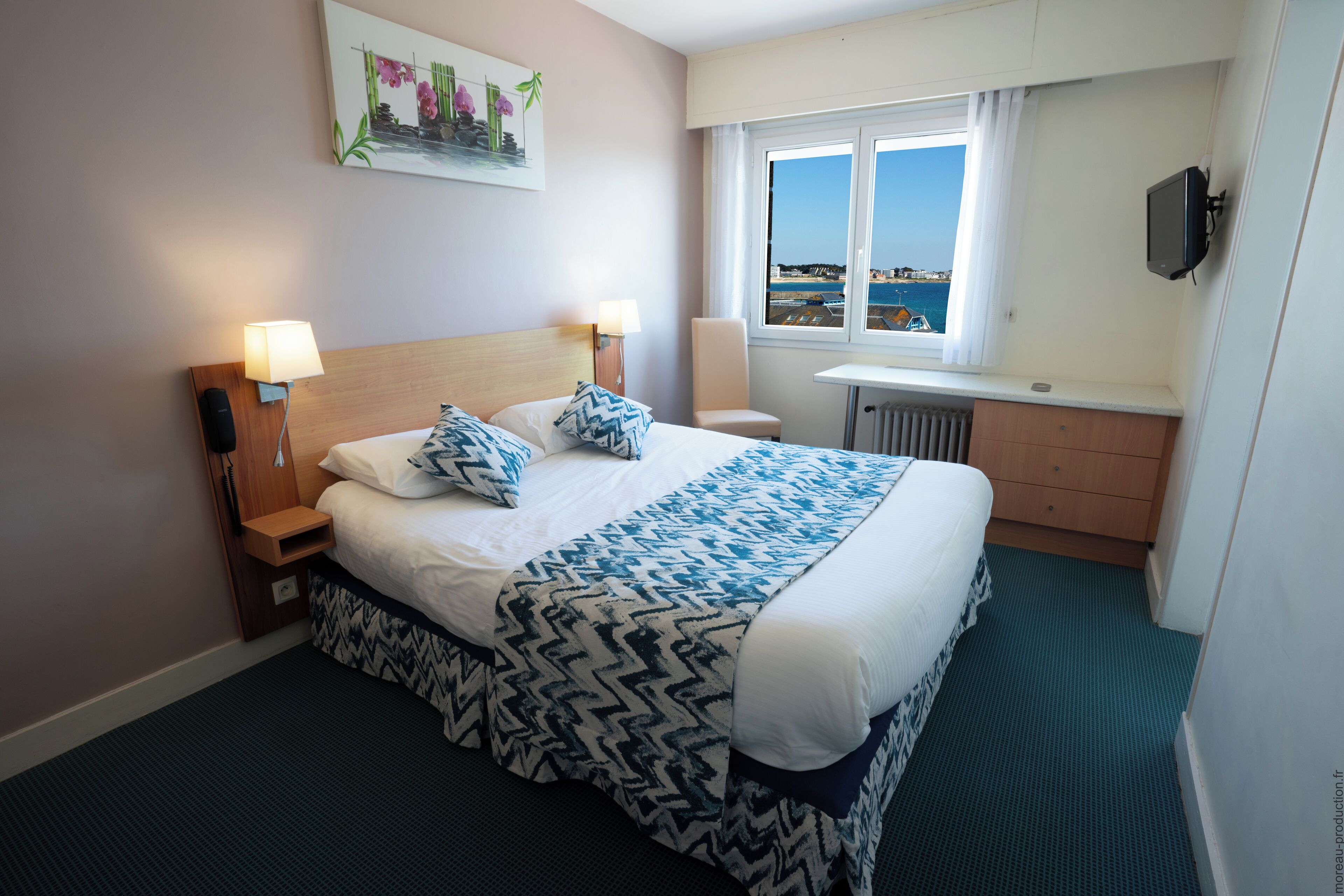 A hotel room with a large bed, two bedside lamps, a view of the sea, a small table, and a TV.