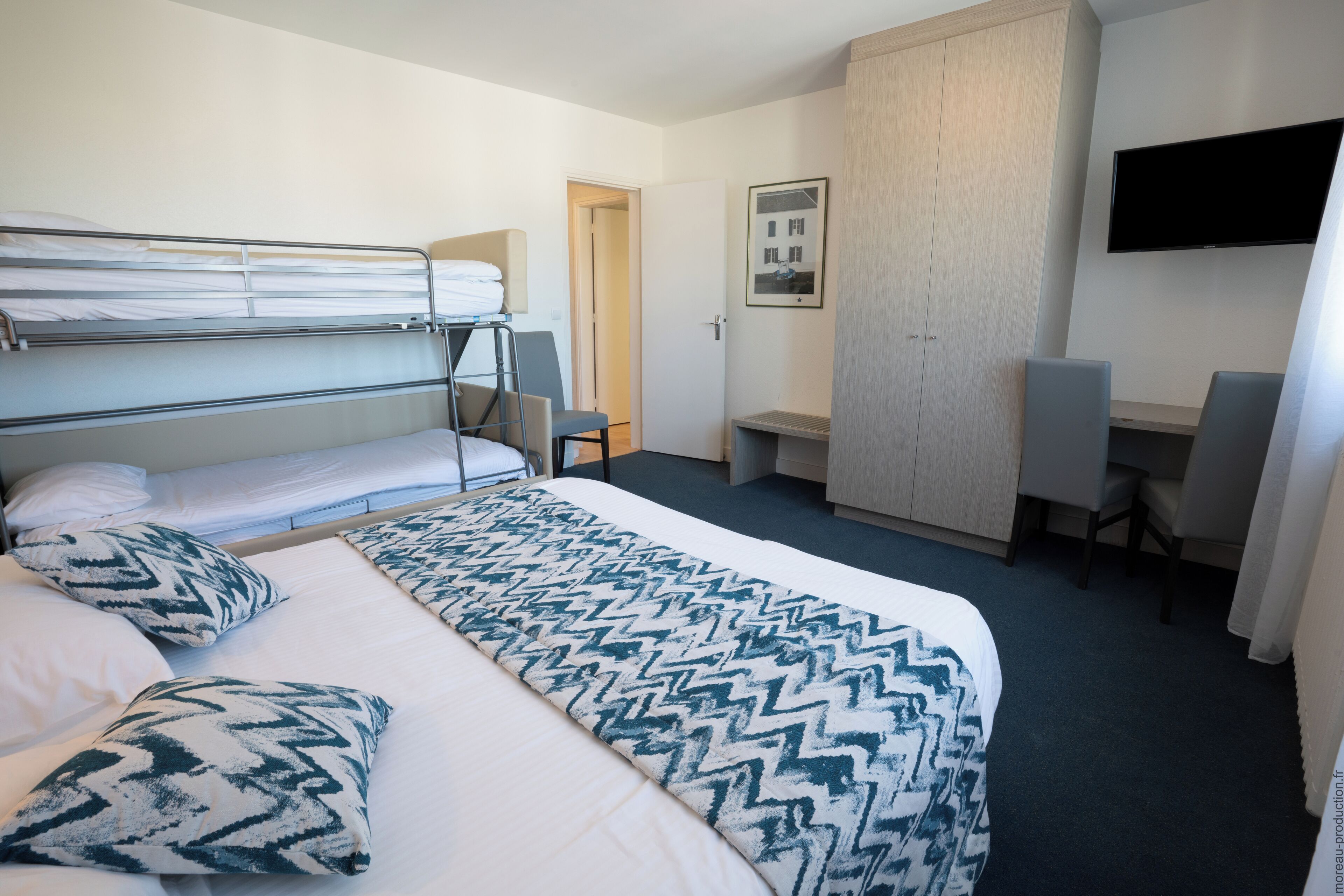 A hotel room with a bunk bed, a desk, a chair, a TV, and a wardrobe.