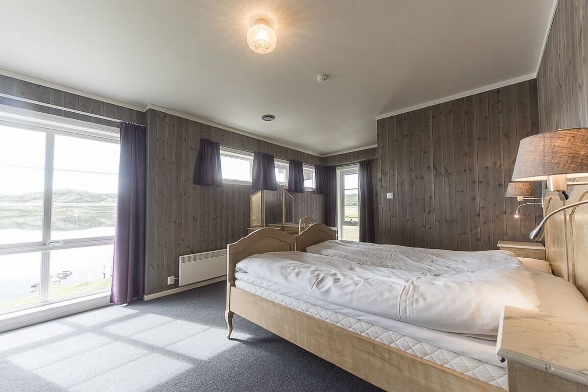 A bedroom with a large bed, wooden walls, a window with curtains, and a bedside lamp.