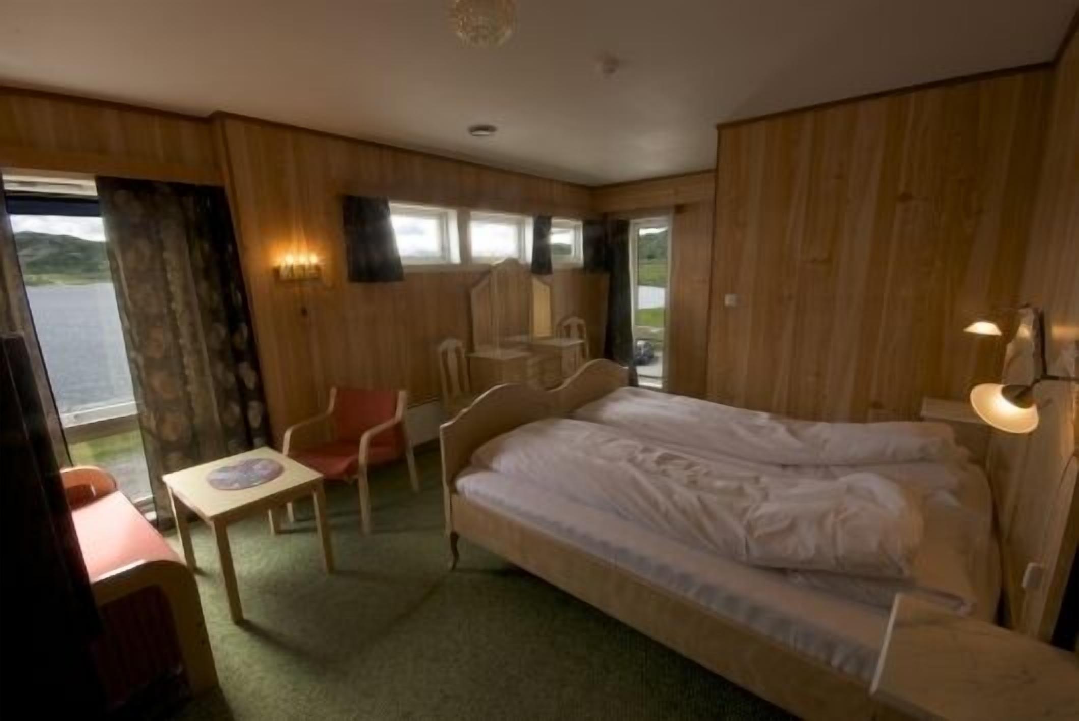 A single-bed room with a wooden headboard, a small table, a chair, and a window with a view of the outdoors.