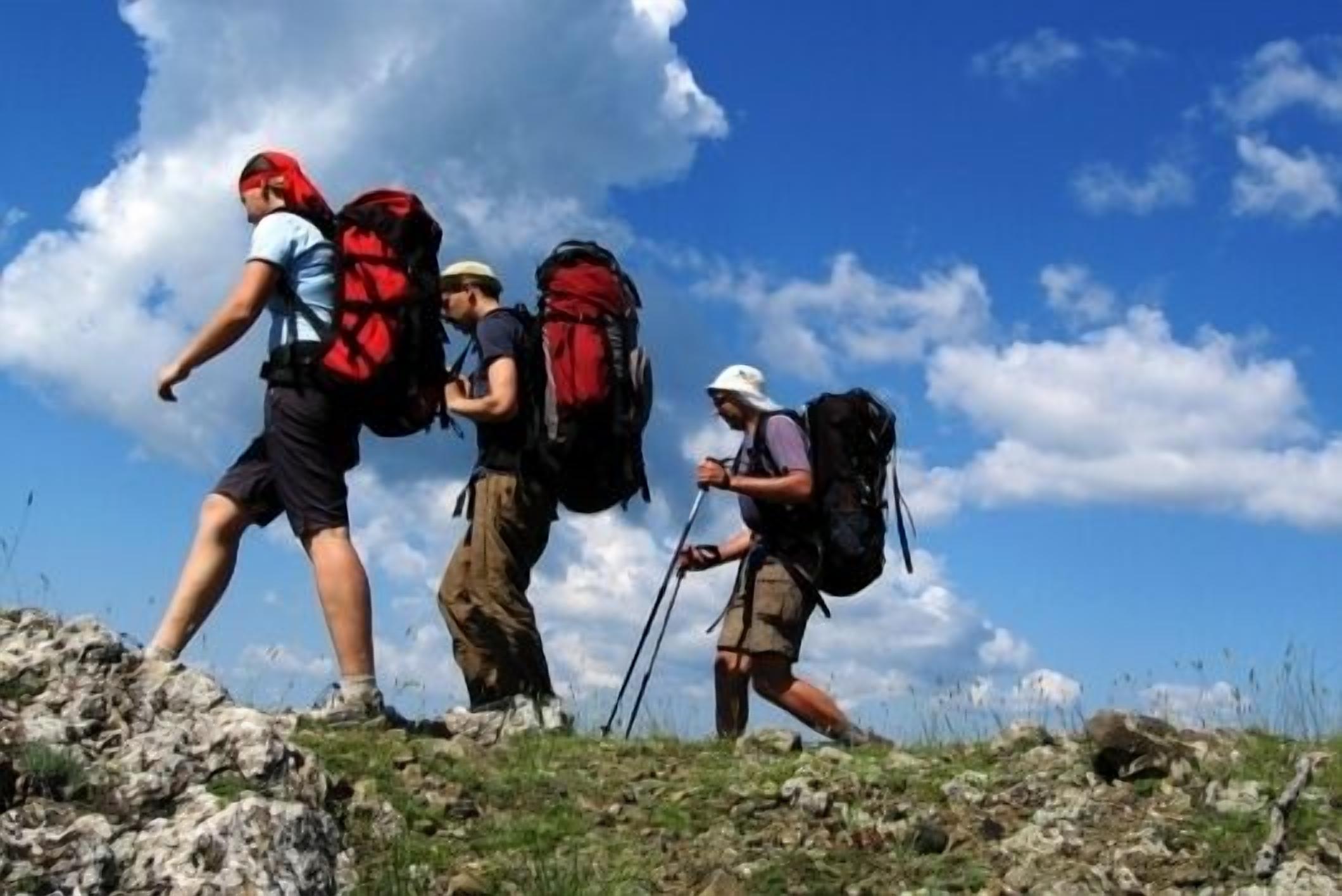 Three hikers on a rocky trail with backpacks, walking under a clear sky.