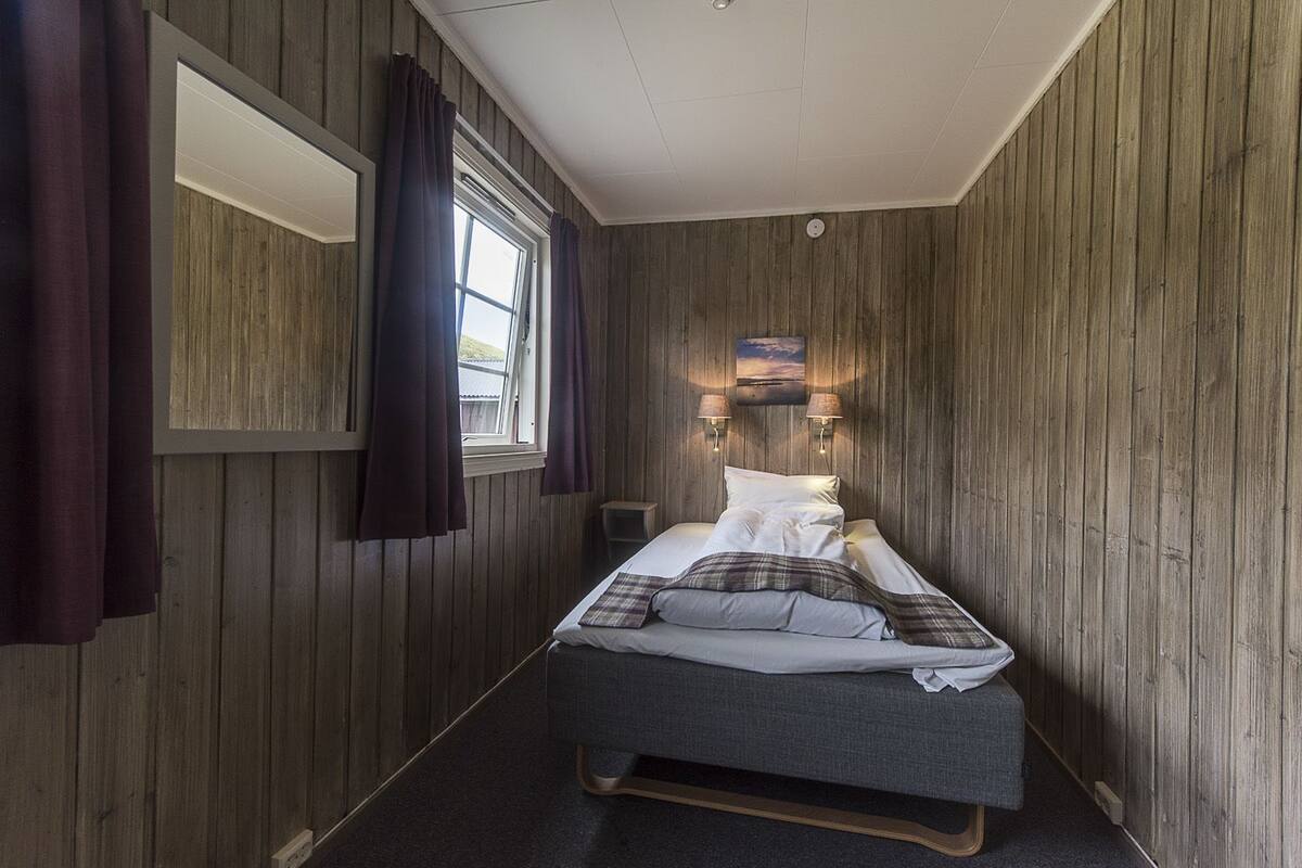 A small, cozy room with wooden walls, a single bed, a window with curtains, and a small table.