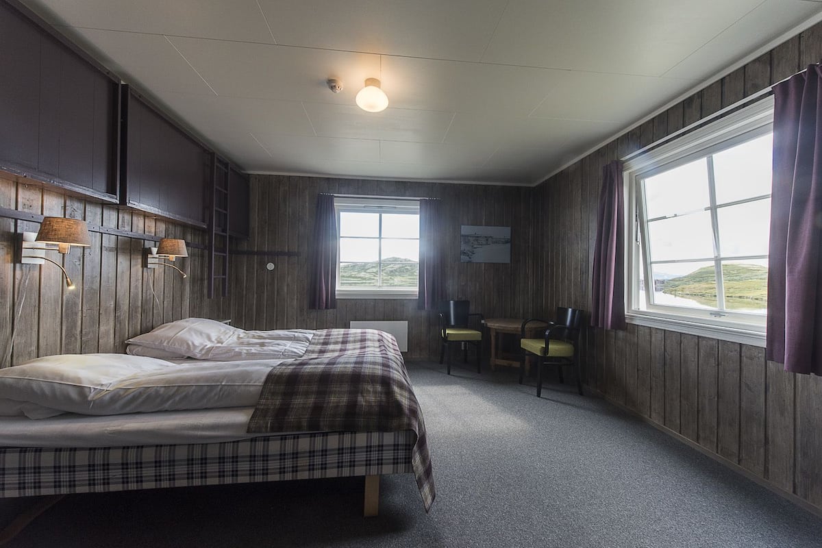 A room with two beds, a table with chairs, a window with curtains, and a wall-mounted light fixture.
