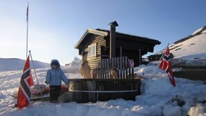A wooden cabin with a deck, surrounded by snow, and flags on either side. - Bergsjøstølen (Al)
