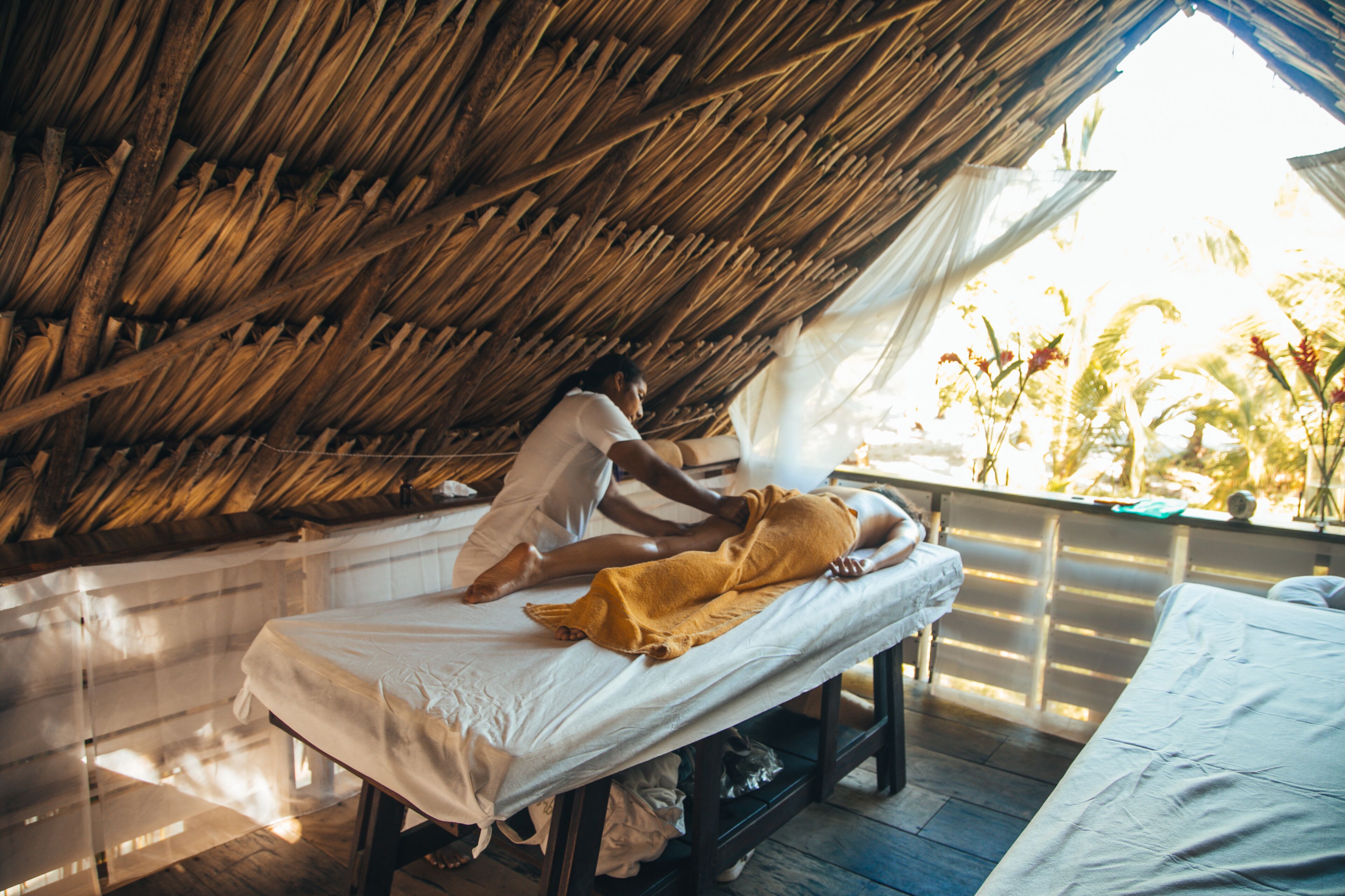 A room with a thatched roof, a bed with a white sheet, and a person lying on the bed.