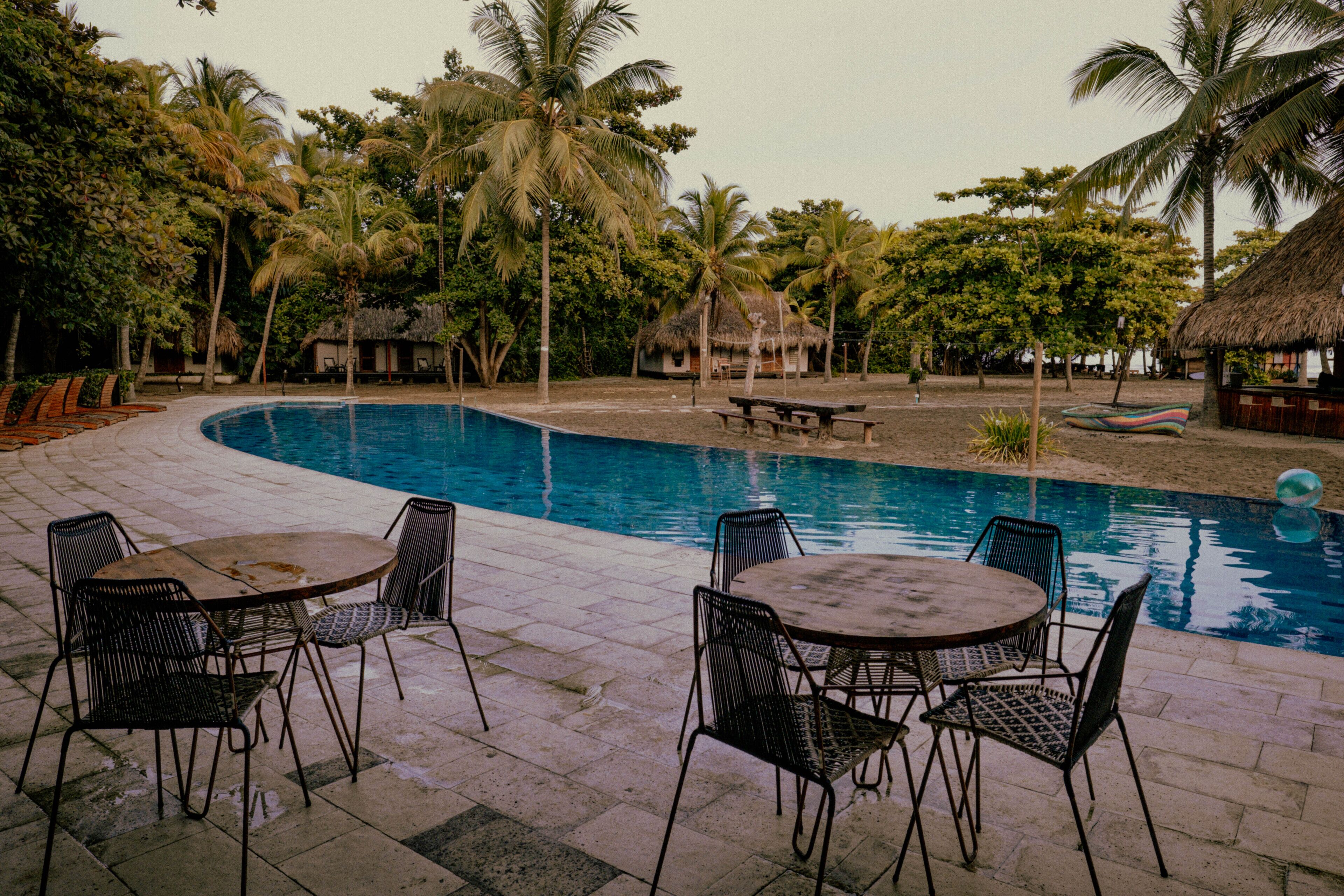 A pool area with lounge chairs and tables, surrounded by palm trees and thatched-roof huts.
