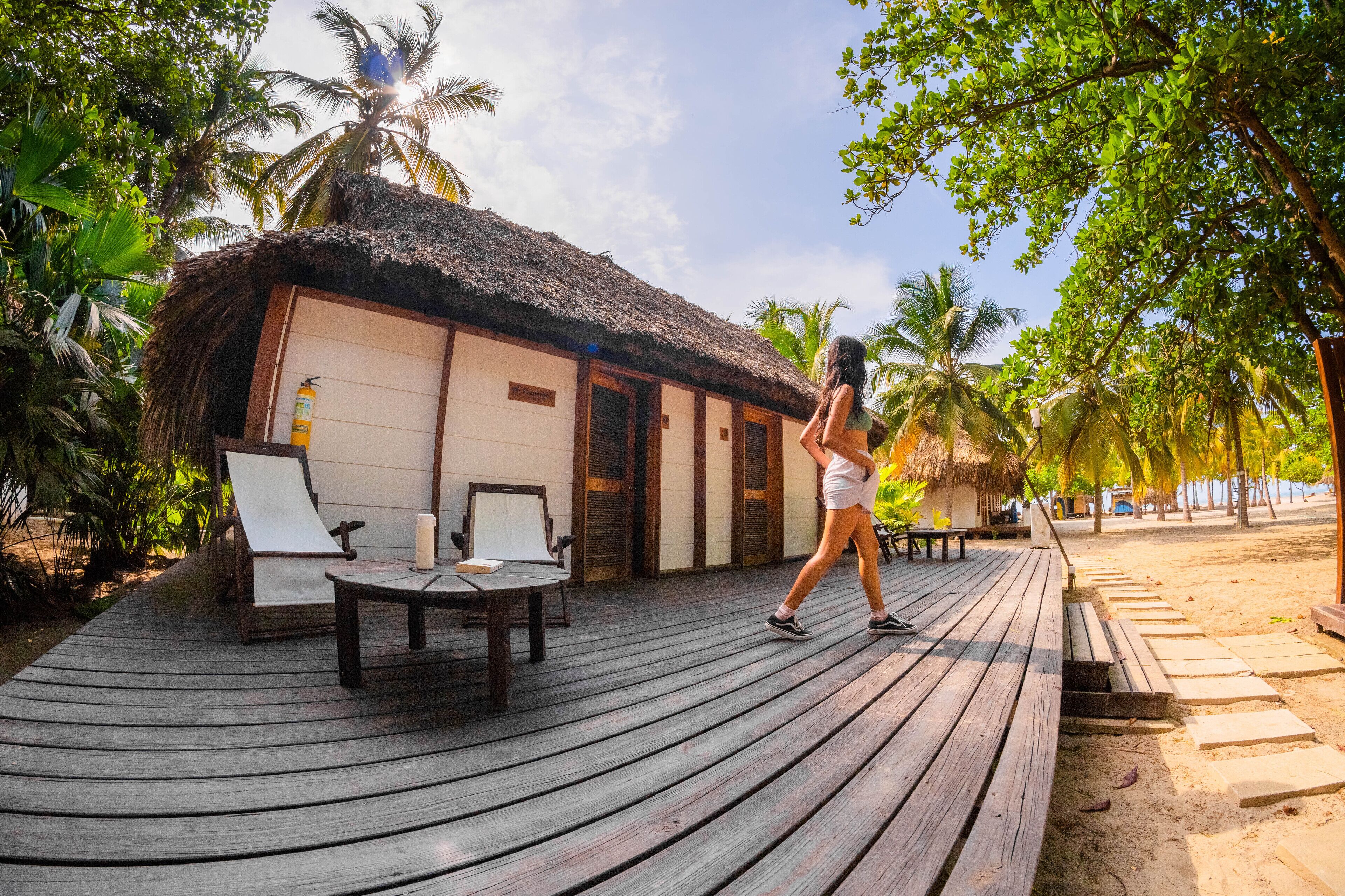 Een houten terras met loungestoelen en een gebouw met rieten dak, waarschijnlijk een resort of hotel.