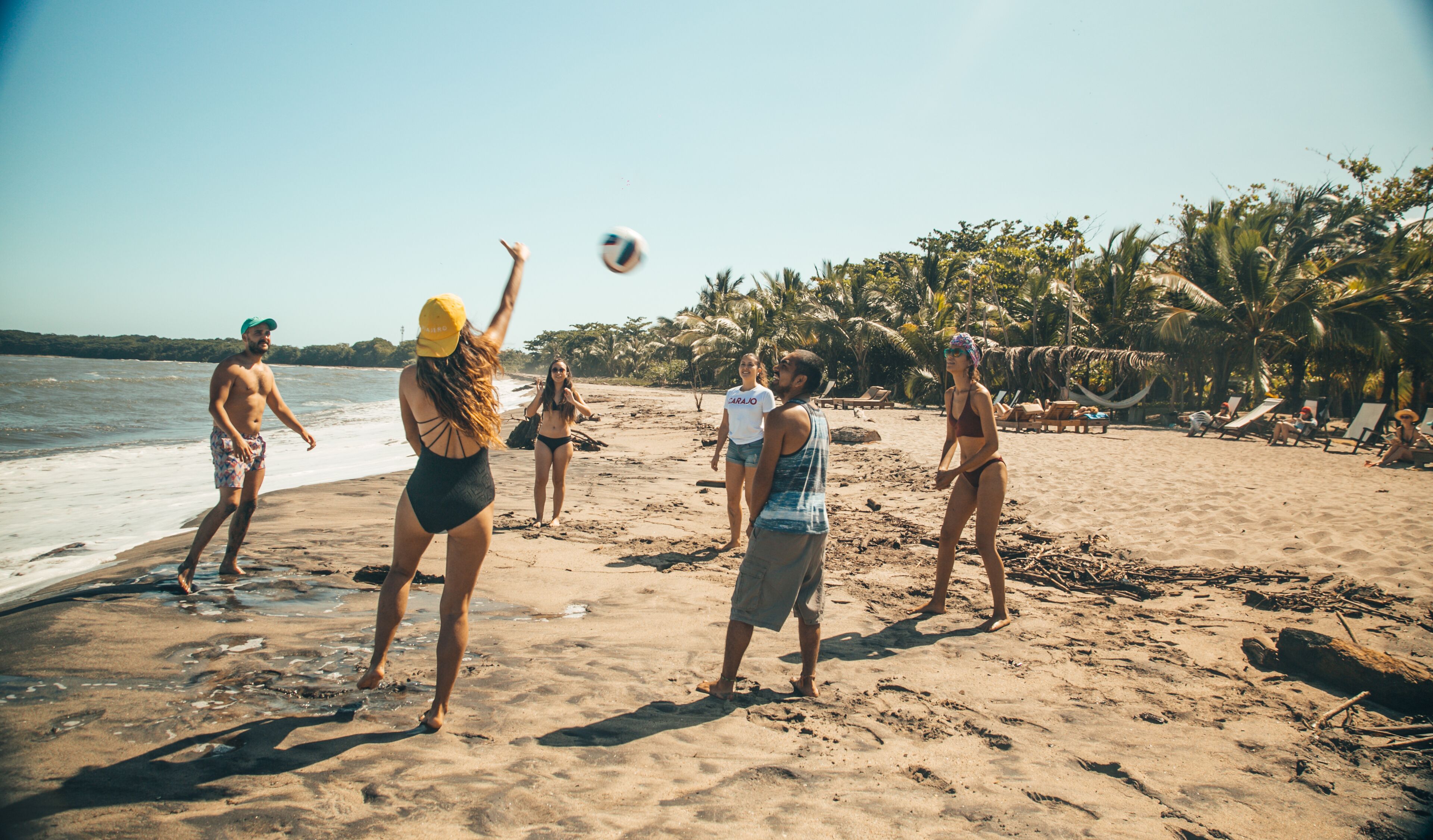 A beach scene with people playing volleyball, palm trees, and beach chairs in the background.