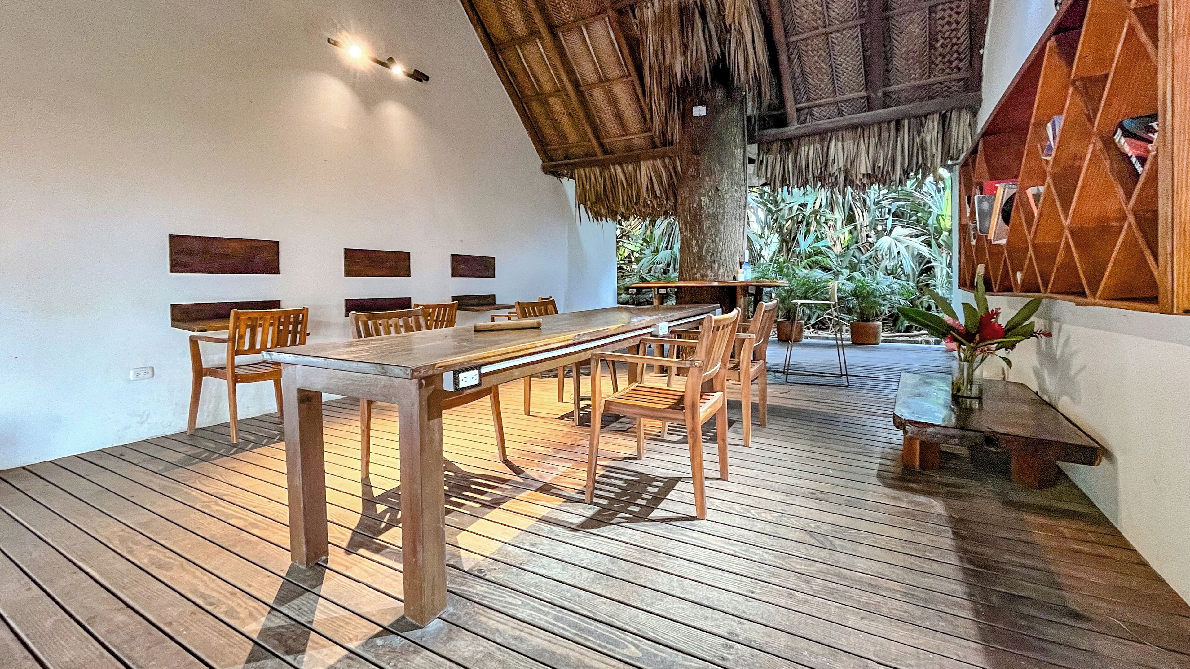 Una terraza de madera con una larga mesa de comedor y sillas, rodeada de plantas en macetas y una estructura de techo de paja.