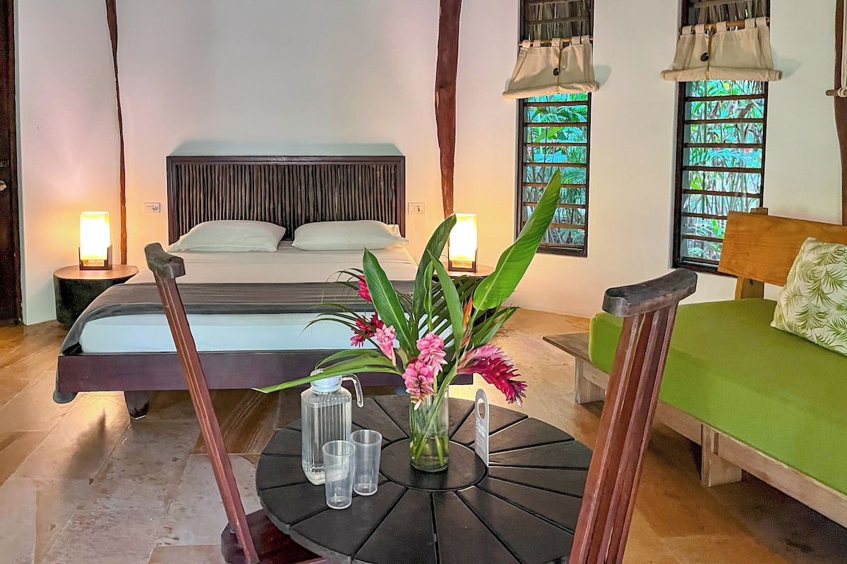 Une chambre d’hôtel avec un lit, une table à manger avec des chaises, un vase de fleurs et deux fenêtres avec des rideaux.