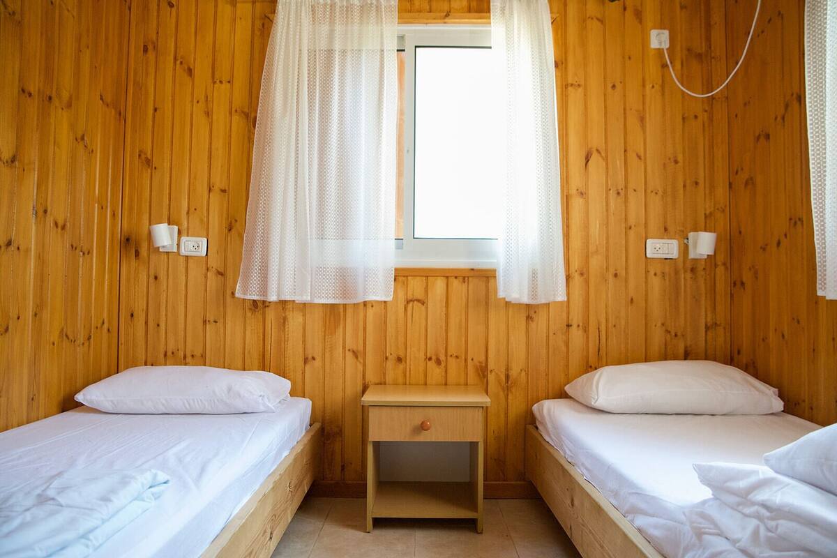 A wooden room with two beds, a small bedside table, and a window with sheer curtains.
