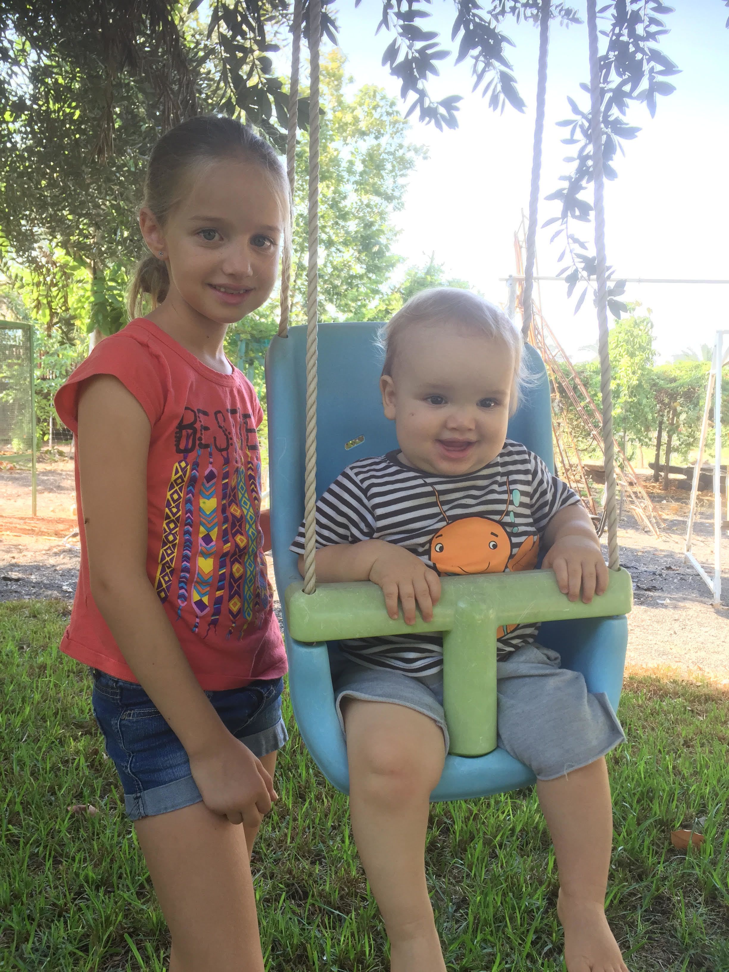 A child in a swing chair with a toy, and another child standing beside it.