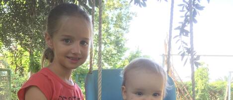 A child in a swing chair with a toy, and another child standing beside it.