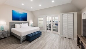 Un dormitorio con cama, mesitas de noche, un banco, televisor, un cuadro y vistas a través de un ventanal.