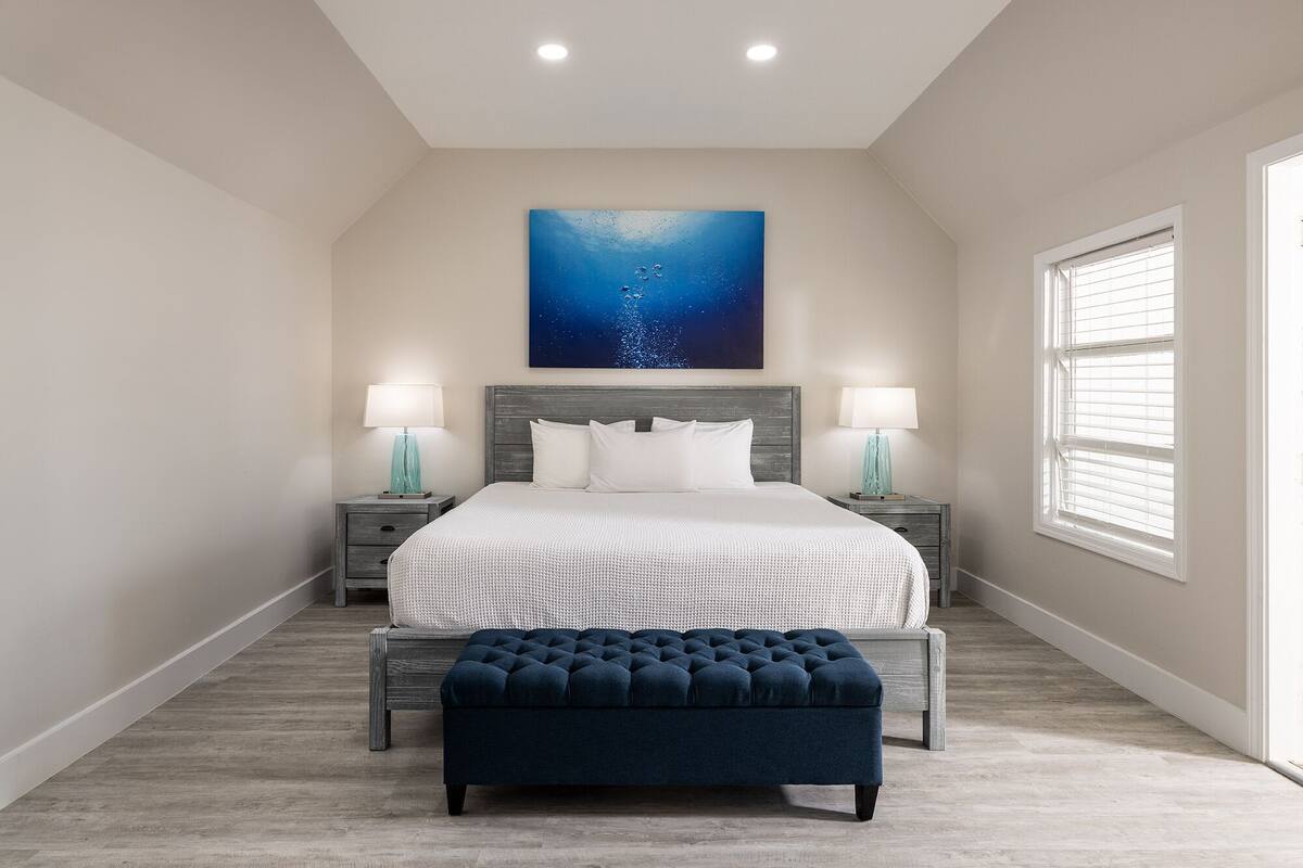 Une chambre avec un grand lit, deux tables de chevet avec des lampes, un pouf bleu, un luminaire au plafond et un grand tableau accroché au mur.