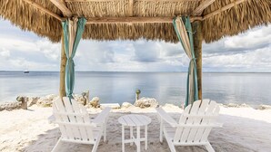 Un espacio de estar junto a la playa con dos sillas Adirondack blancas y una mesita bajo una estructura de techo de paja.