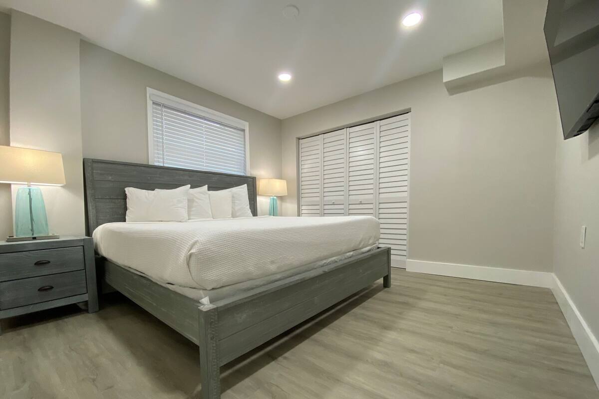 Une chambre à coucher comprenant un lit, des tables de chevet, une fenêtre avec des stores, un placard avec des portes à persiennes et un téléviseur à écran plat fixé au mur.