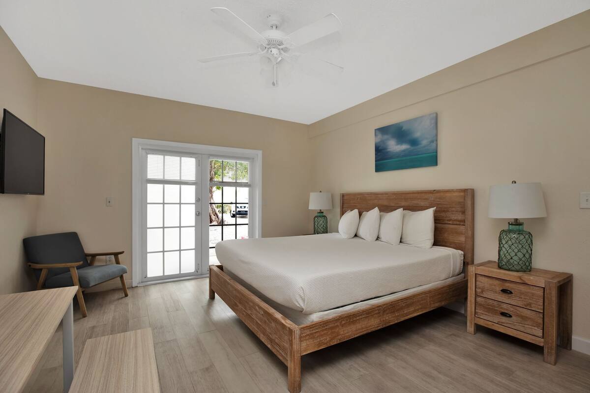 Une chambre à coucher avec un lit en bois, deux tables de chevet avec des lampes, une chaise, un téléviseur et une grande fenêtre avec des stores.