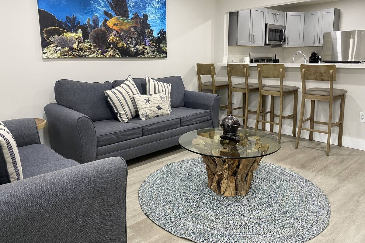 Un salon moderne avec un canapé gris, des coussins à rayures et une table basse en verre avec un socle en bois. Un grand aquarium contenant des coraux et des poissons est fixé au mur.