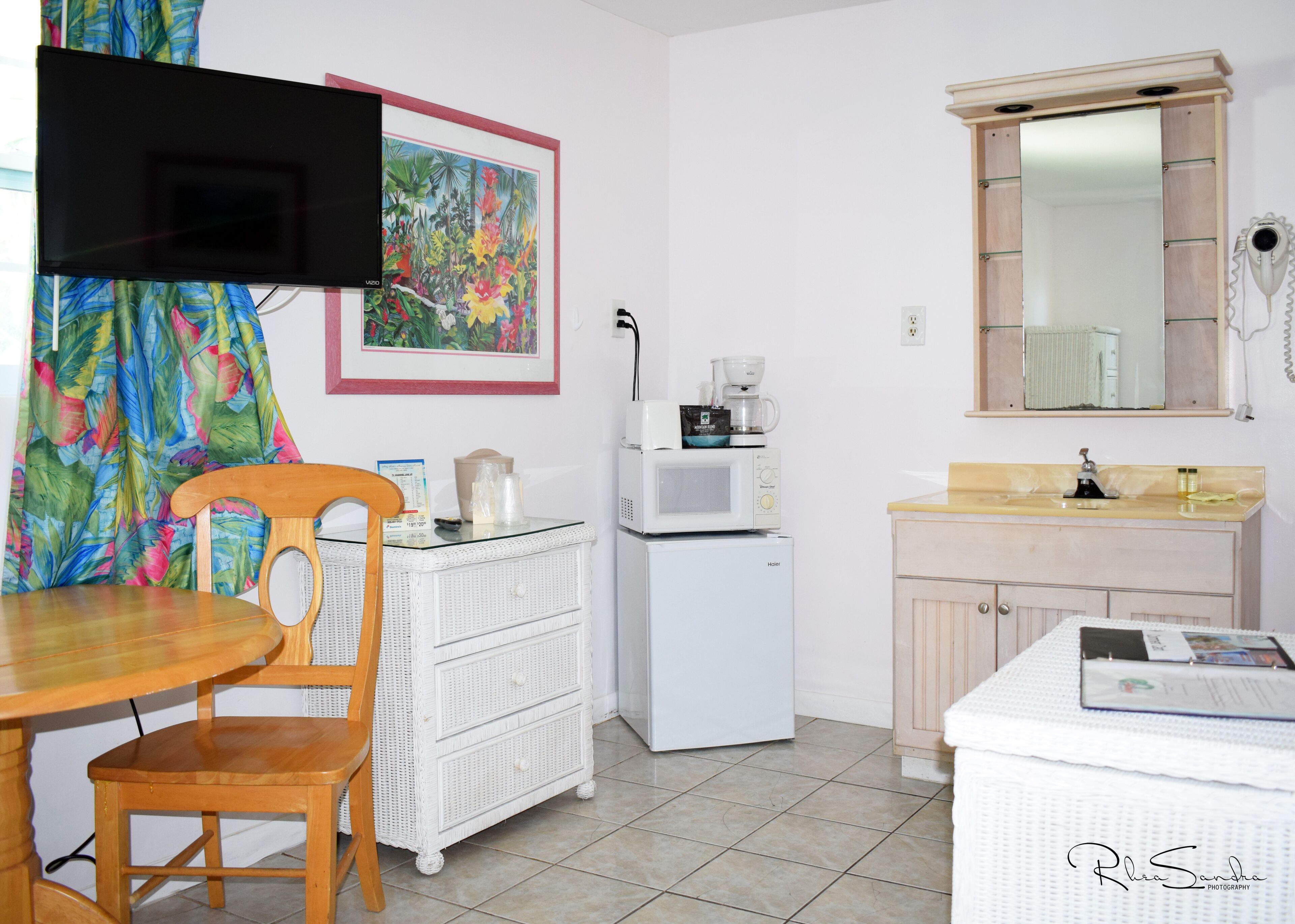 A compact kitchen with a microwave, coffee maker, and a small refrigerator. There is a dining table with a chair, a white wicker chest of drawers, and a mirror above the sink. A colorful tropical print is visible on the curtain.