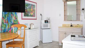 A compact kitchen with a microwave, coffee maker, and a small refrigerator. There is a dining table with a chair, a white wicker chest of drawers, and a mirror above the sink. A colorful tropical print is visible on the curtain.