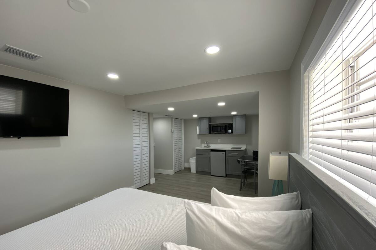 Une chambre d’hôtel moderne équipée d’un lit, d’une télévision, d’une kitchenette et d’une fenêtre avec des stores.