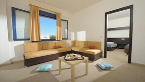 A modern living room with a sofa set, a coffee table with breakfast items, and a view through a window with curtains. - Meropi Hotel & Apartments (Hersonissos)