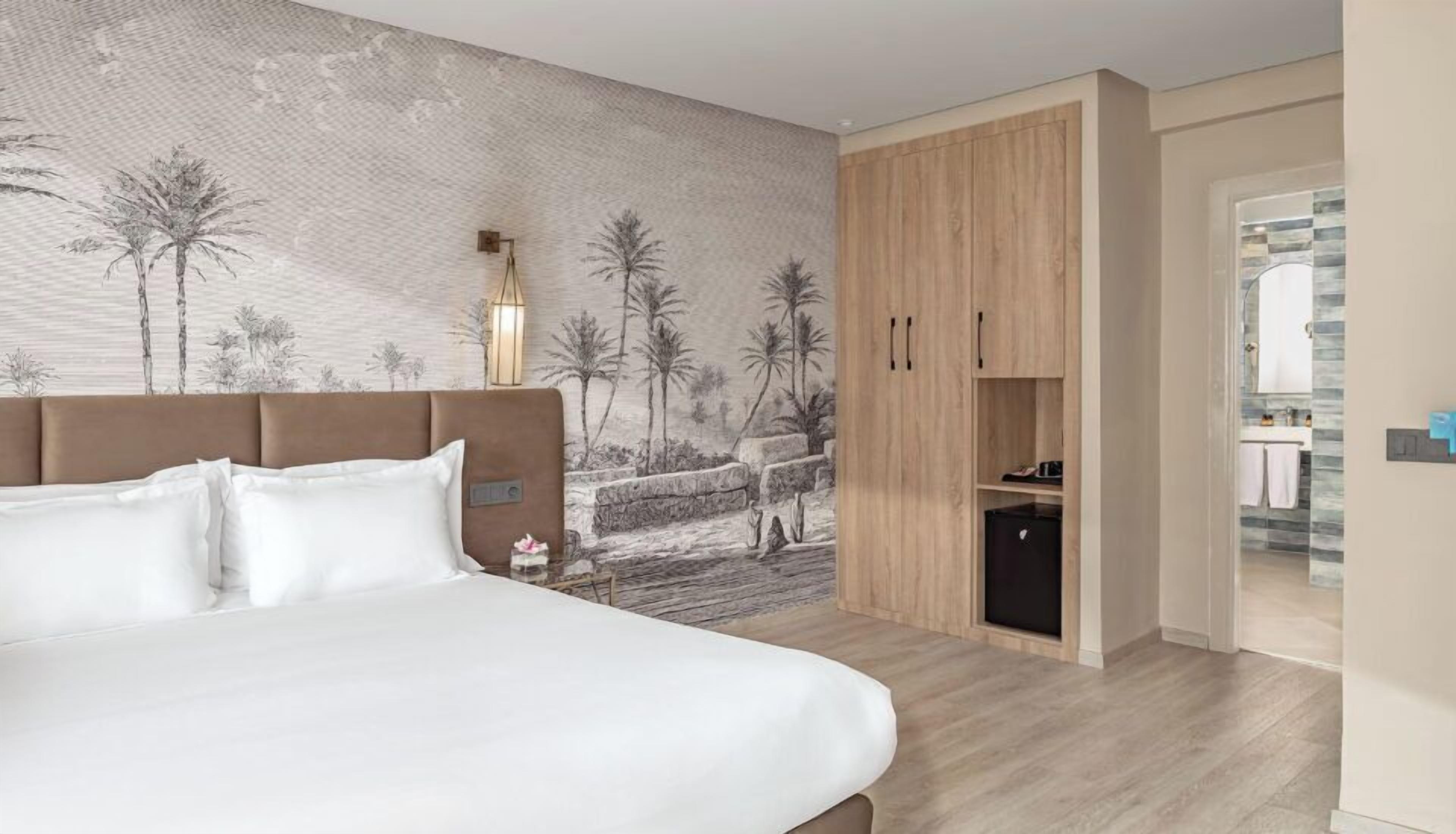 Une chambre à coucher comprenant un lit, une armoire, une table de chevet, une fresque murale, et une salle de bain visible à travers une porte ouverte.