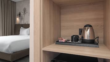 Quarto de hotel com cama, pequeno espaço de cozinha e mesa de cabeceira com cafeteira e bandeja de objetos.