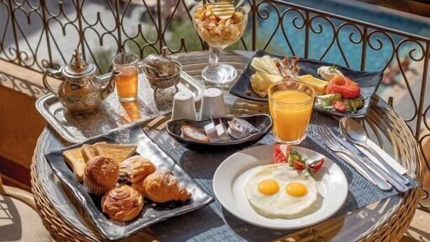 A dining table with breakfast items including croissants, eggs, and a fruit salad, set on a balcony with a pool view. - Grand Plaza (Marrakech)