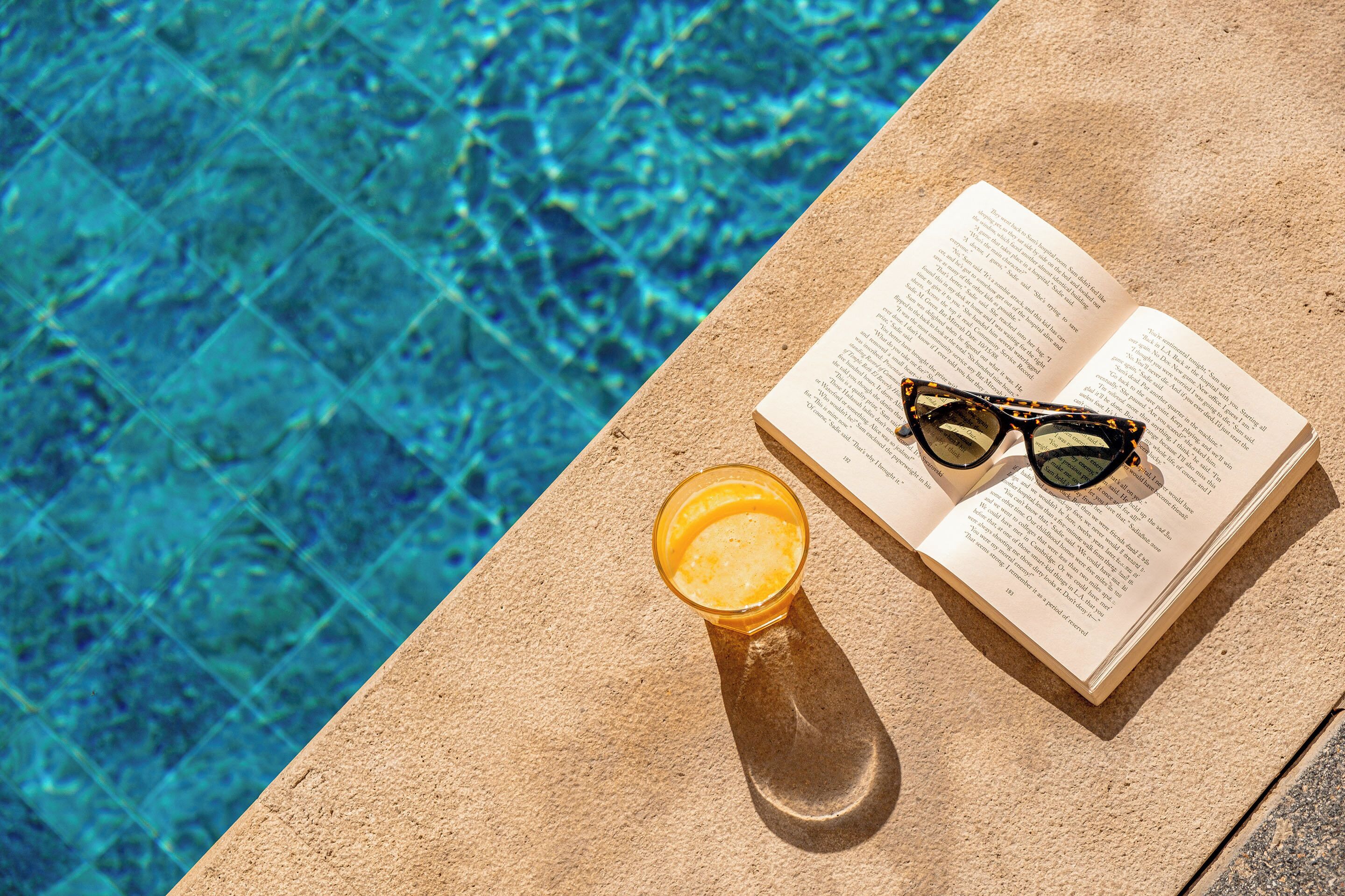 An open book with glasses and a drink on a concrete surface by a pool.