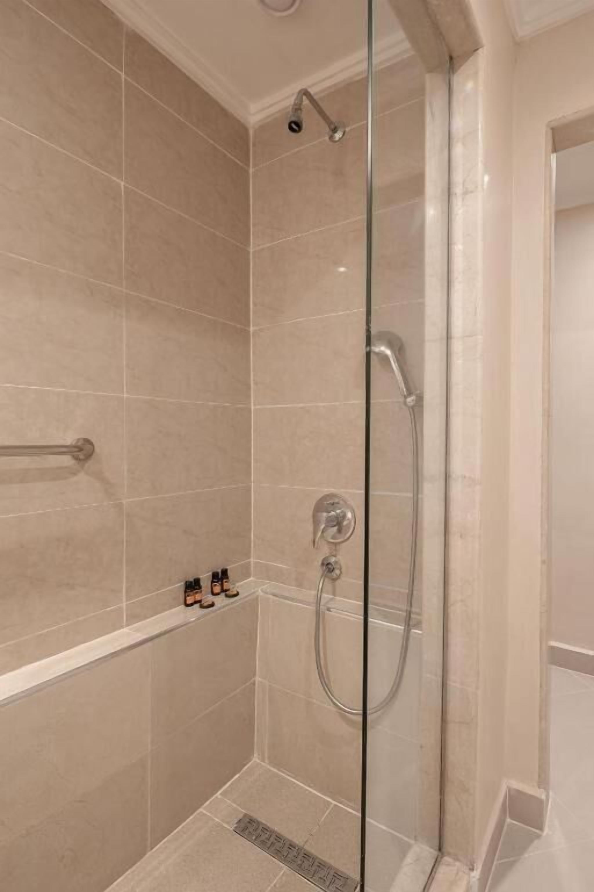 A modern bathroom with a glass-enclosed shower, beige tiles, and a small shelf with toiletries.