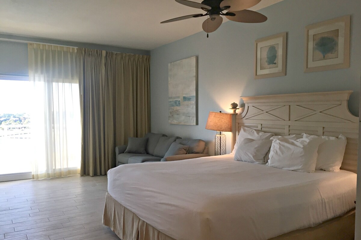 A bedroom with a large bed, a ceiling fan, a nightstand with a lamp, a couch, and a window with curtains.
