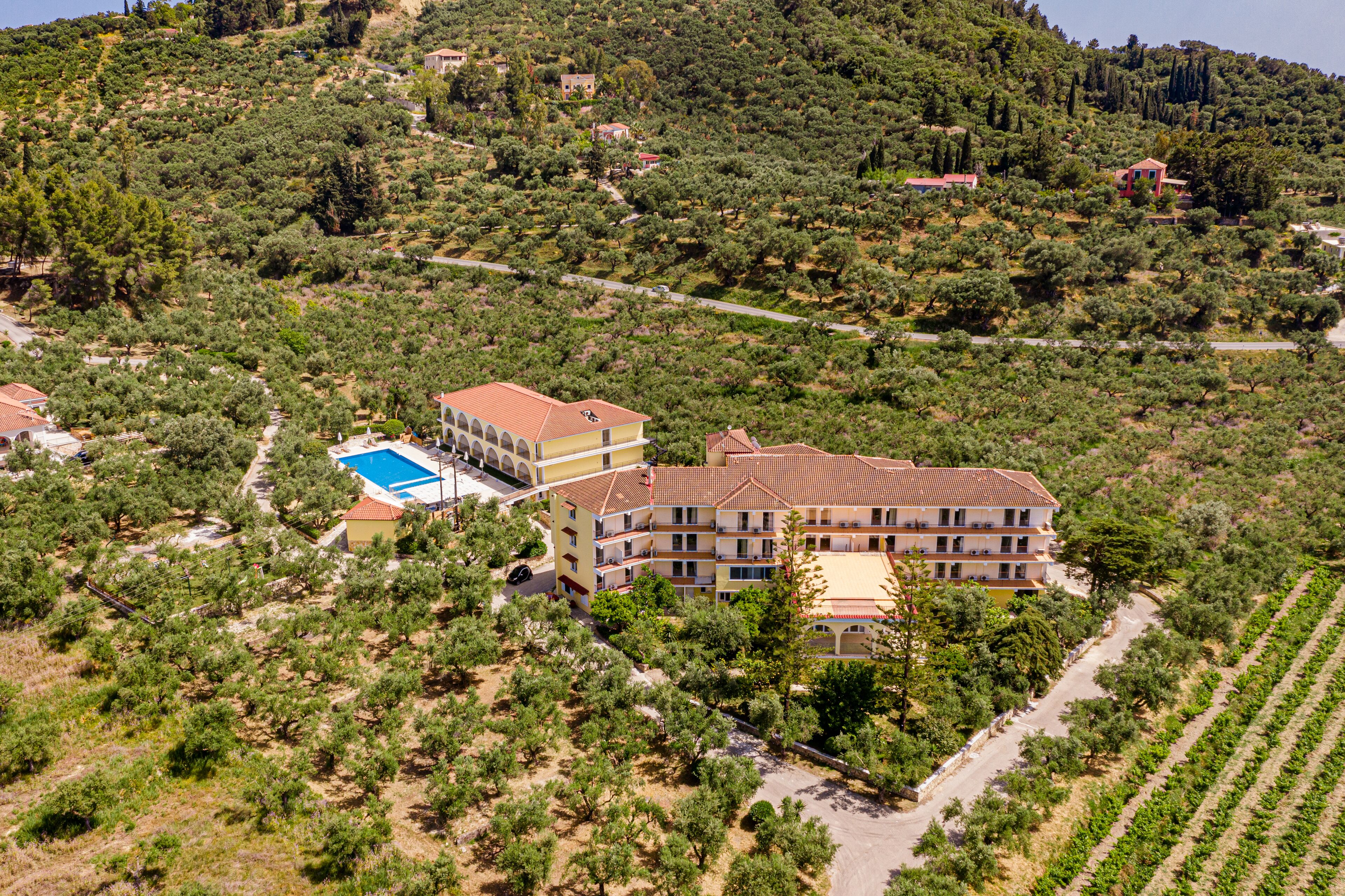 Un hotel grande de varios pisos con tejado de tejas rojas, rodeado de olivares y un paisaje montañoso.