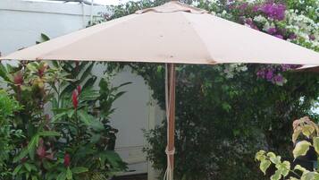 Un espace extérieur aménagé avec une table et deux chaises, protégé par un grand parasol, et entouré d’une végétation luxuriante et de plantes en fleurs.