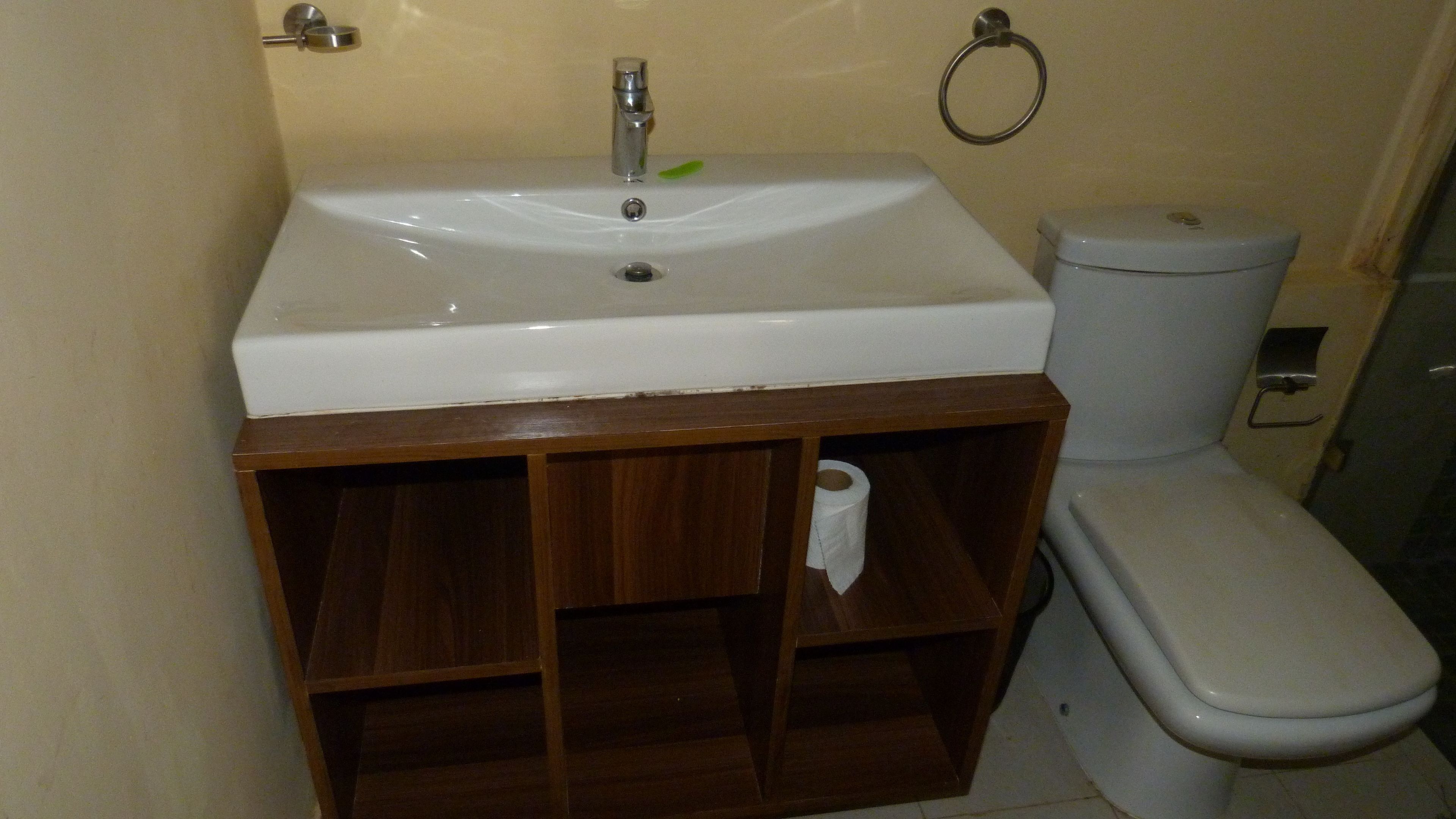Un baño con lavamanos blanco, tocador de madera, inodoro y un perchero para toallas en la pared.