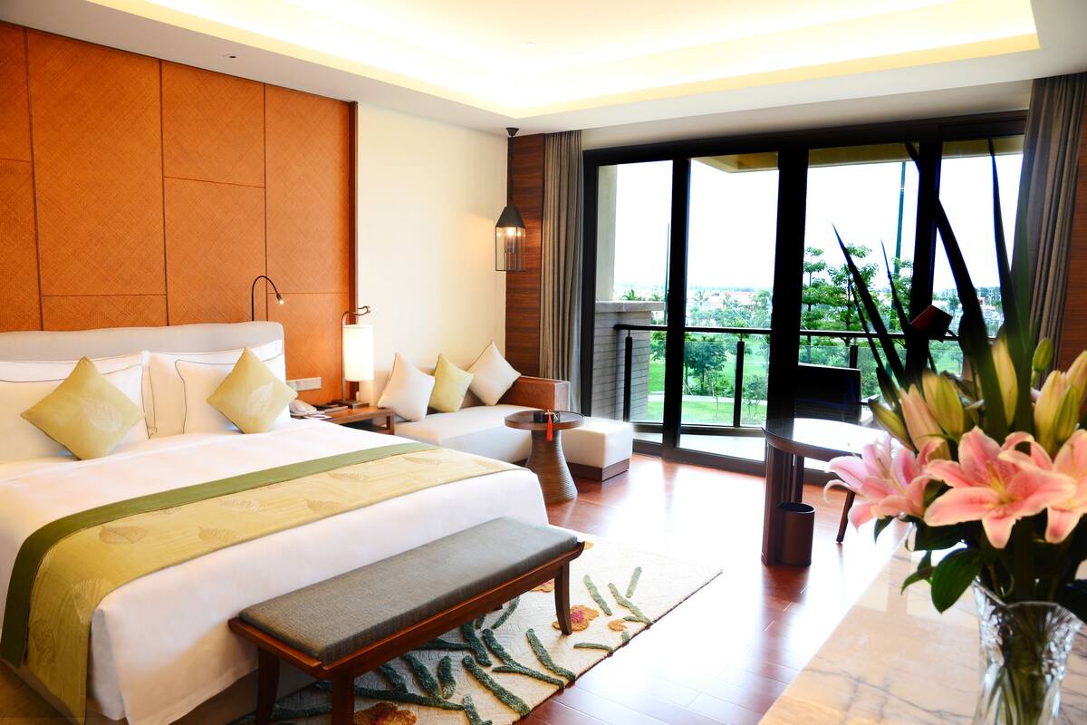 Une chambre d’hôtel avec un grand lit, une banquette, un bureau, une chaise, une télévision et une vue sur la verdure par les fenêtres.