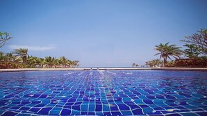 Une piscine d’un bleu éclatant, ornée de motifs en mosaïque, entourée de palmiers et surplombée d’un ciel dégagé.