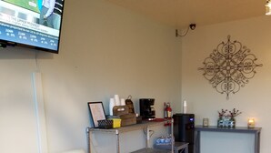 A living room with a light blue armchair, a grey bench, a coffee station, a TV mounted on the wall, and a decorative wall piece. - SureStay by Best Western Yerington (Yerington)